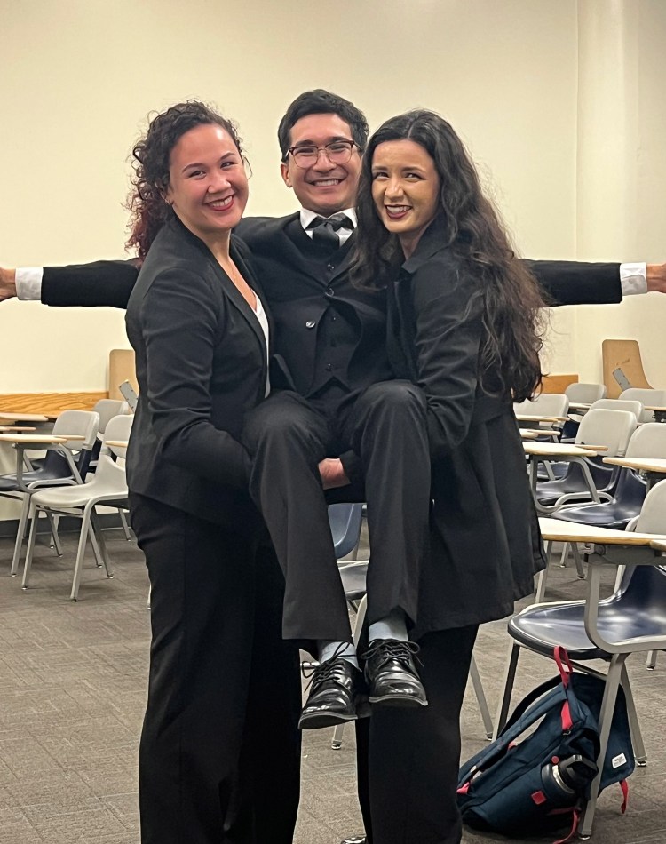 Two women hold up a man with his arms extended out. They all have black suits on.