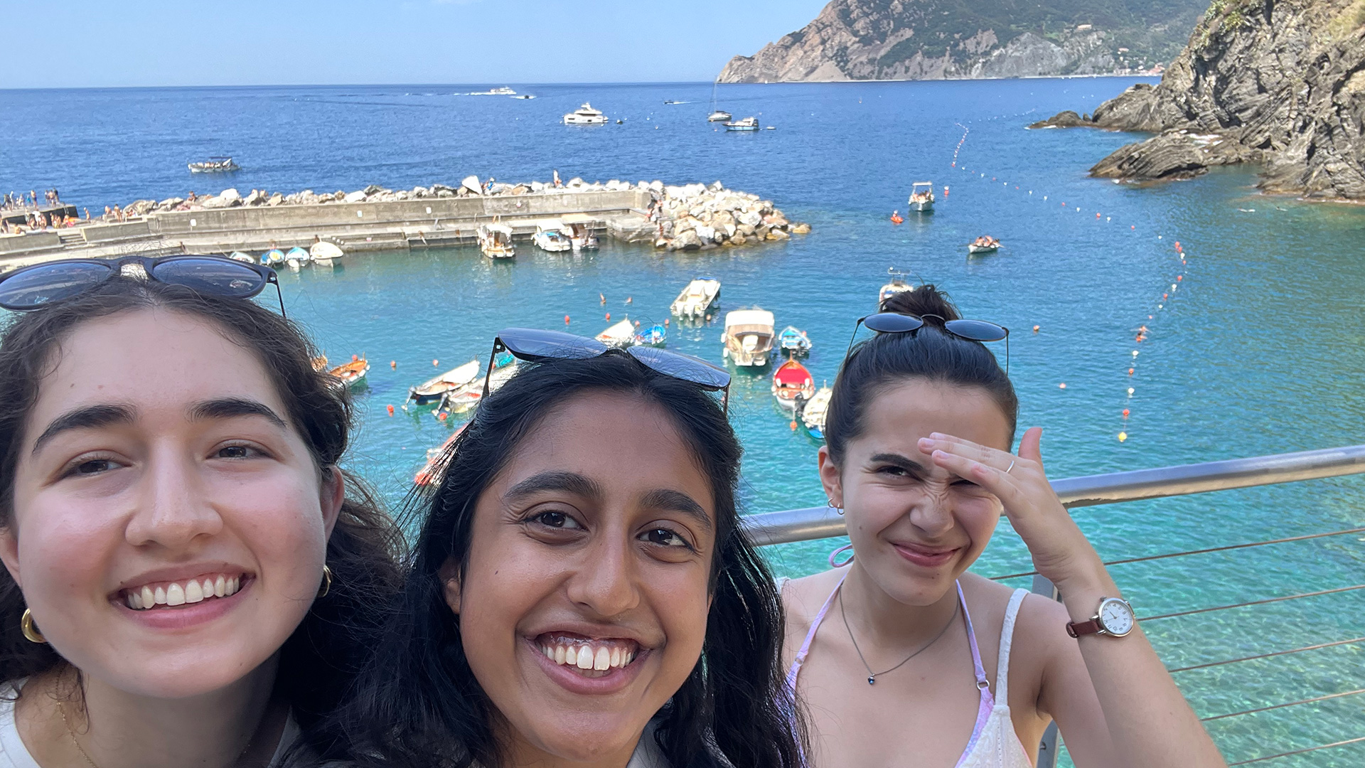 Three women pose in front of the coast. Boats and a dock can be seen in the background, as well as rocky cliffs.