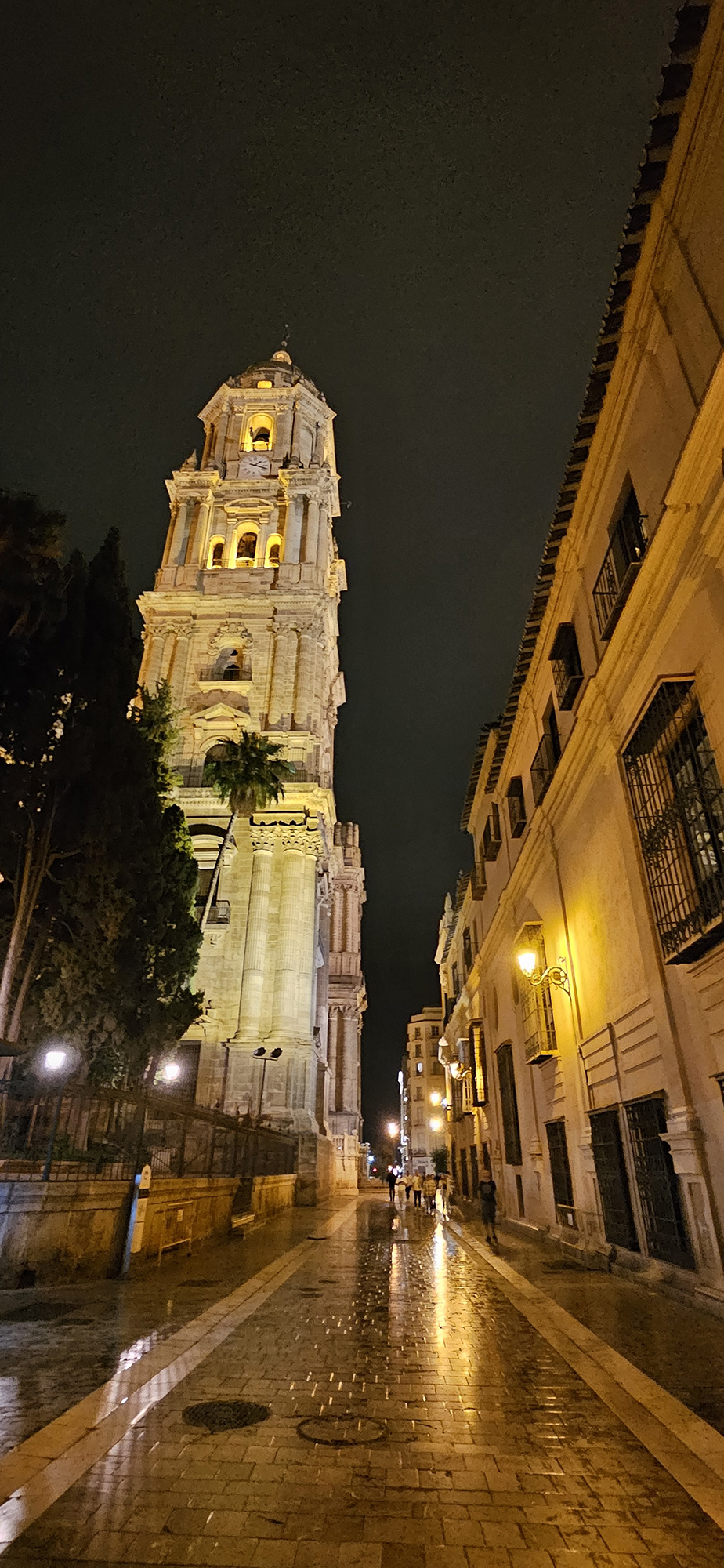 An old street at night with brick roads and old architecture, including a tall tower is lit by lamp posts.