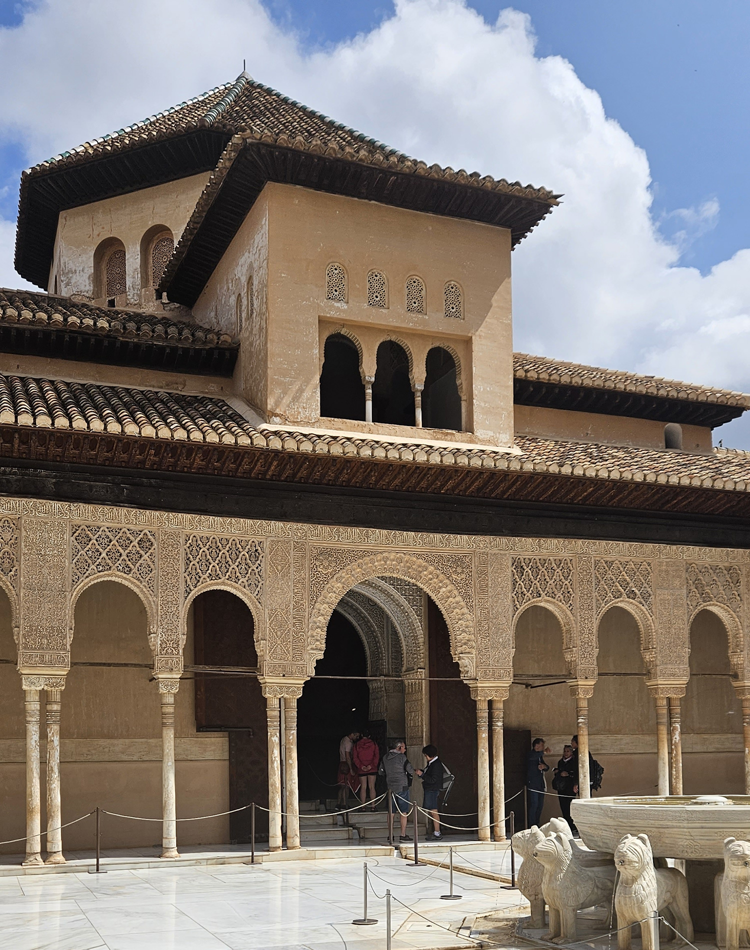 A cream colored old building can be seen with dark brown roofs. There is a fountain on the bottom right with carves animals.