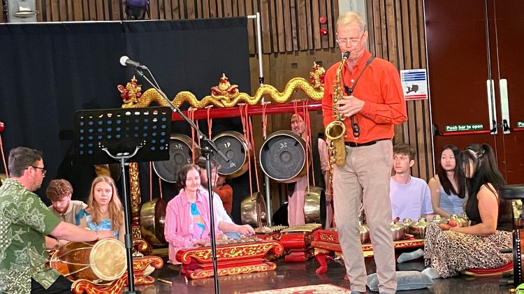 Ben Boone stands playing the saxophone while other musicians sit on the ground and play Indonesian percussion instruments. Ben Boon is in a Orange collard shirt and tan pants. There are is a structure for the gamelan which has golden dragons on it.