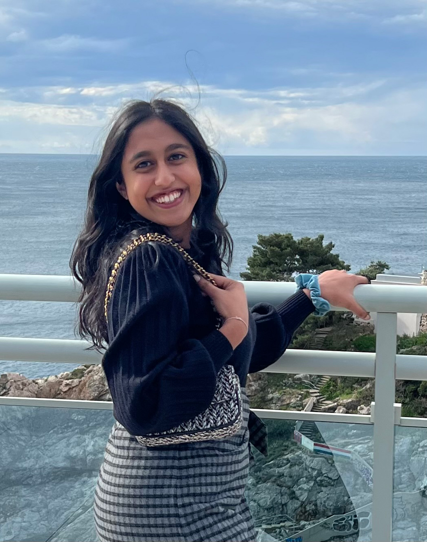 A woman stands on a ledge which overlooks the edge of the coast, with the ocean in the background.