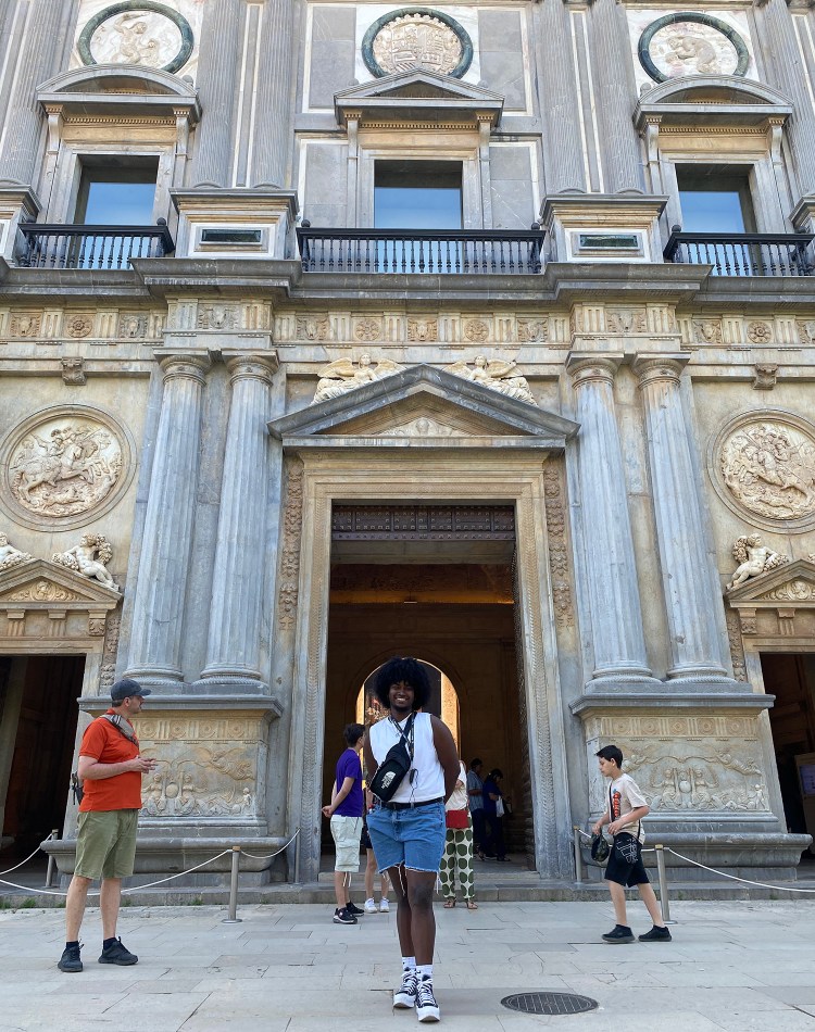 Shaymany Suarez stands in front of a large building in a white sleeveless shirt, jorts, and black shoes.