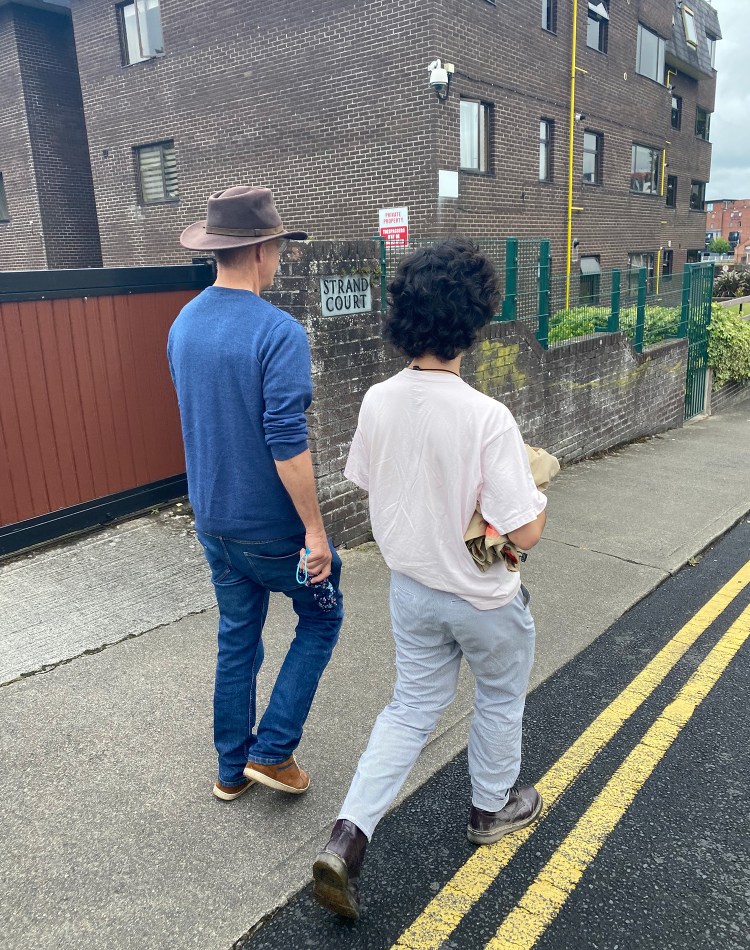 Two men walking down a street. One has a cowboy hat and is wearing all blue, the other has a pink shirt and light blue pants. There is a brick building in the background.