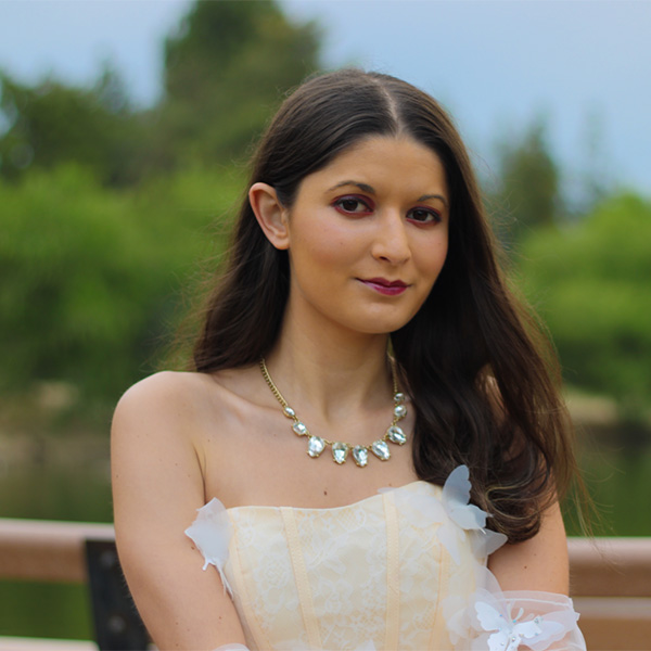 Alexandra Cota in a white formal dress and silver and gold necklace in from of a green backgound of trees.