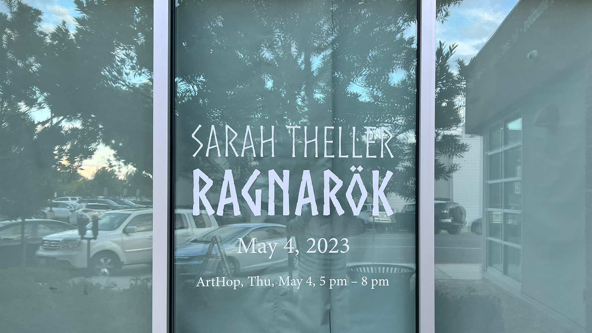 In a window is a display for Theller's show. Her name and show title, "Sarah Theller" and "Ragnarok" are in a font reminiscent of Nordic runes. Below is the date and location.