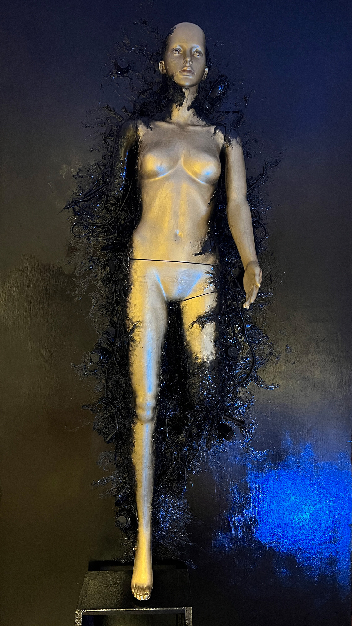 There is a silver mannequin laying of the ground with a missing right arm and left leg. There is a black substance which appears to be consuming the mannequin, or pulling it into the floor.