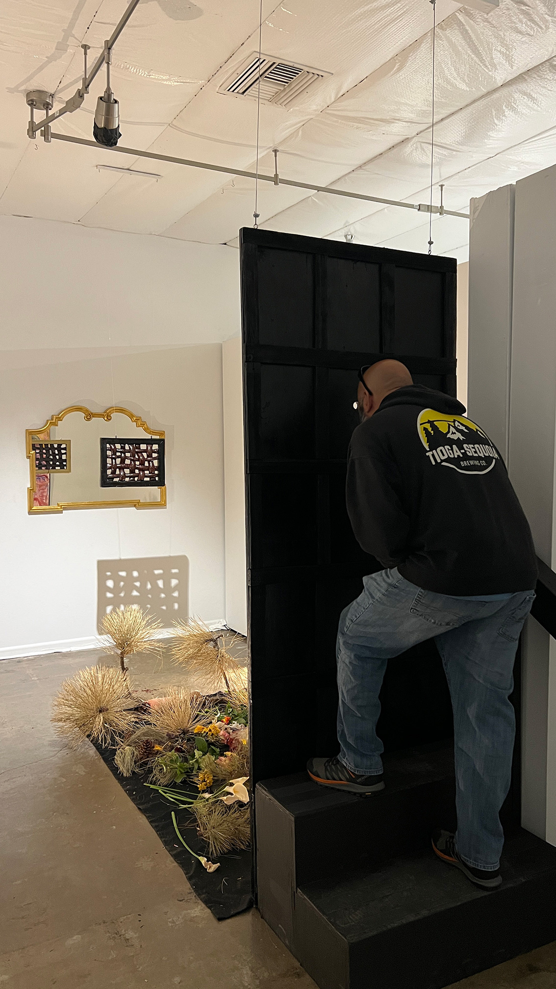 There is a man standing on steps which allow him to look through a peep hole. There is a mirror on the opposite wall that will reflect the art piece back for the person looking through the peephole. We can see dead pine branches, flowers, food, etc. on the ground in front of the piece.