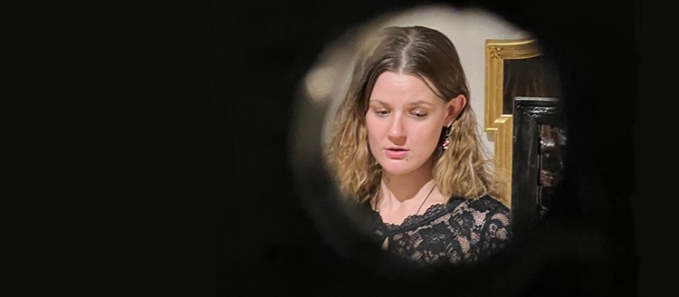 A woman can be seen through a circle looking down. She is in front of a mirror and has brown and blonde hair and a black dress.