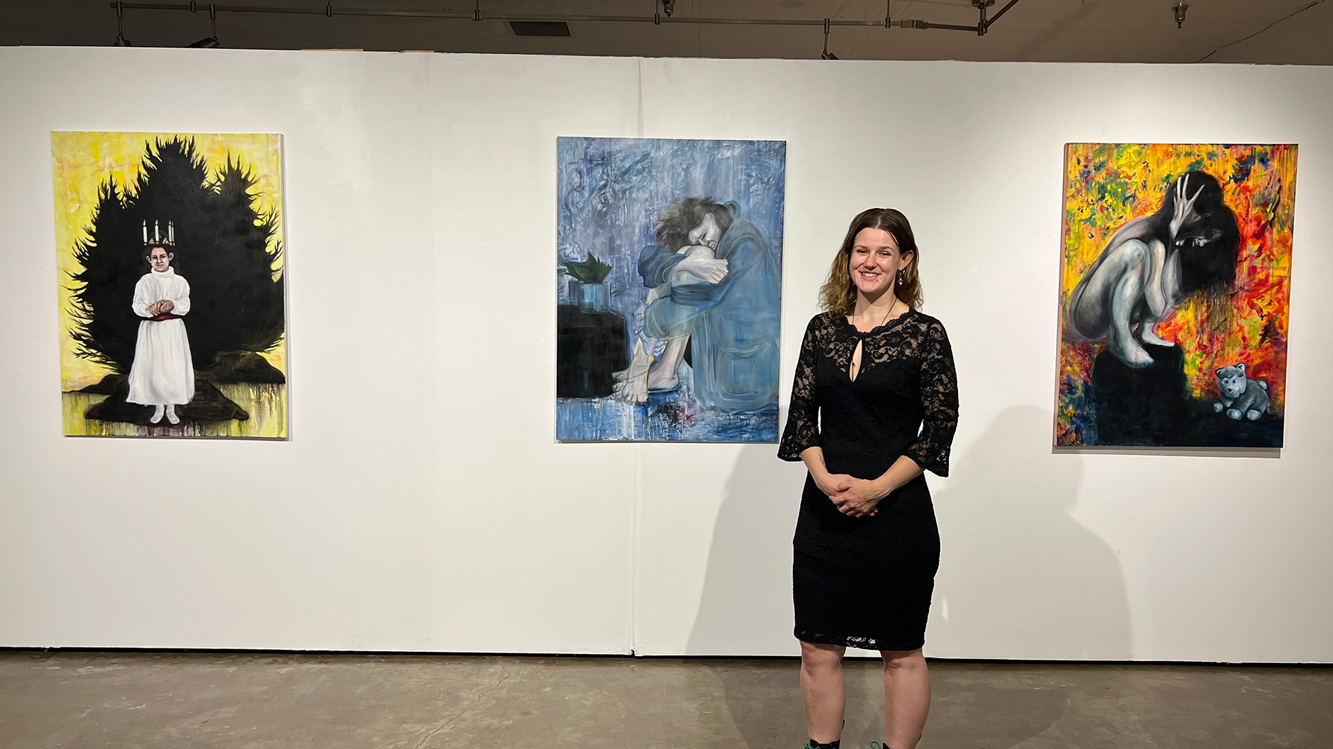 A woman stands in front of a white wall with three paintings on it. She is in a black dress with lace and has brown hair. The paintings represent different elements of death and rebirth.