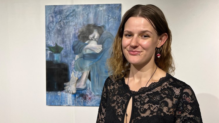 A woman stands in front of an artwork of a woman hugging her knees to her chest. The color blue is the most prominent in the piece.
