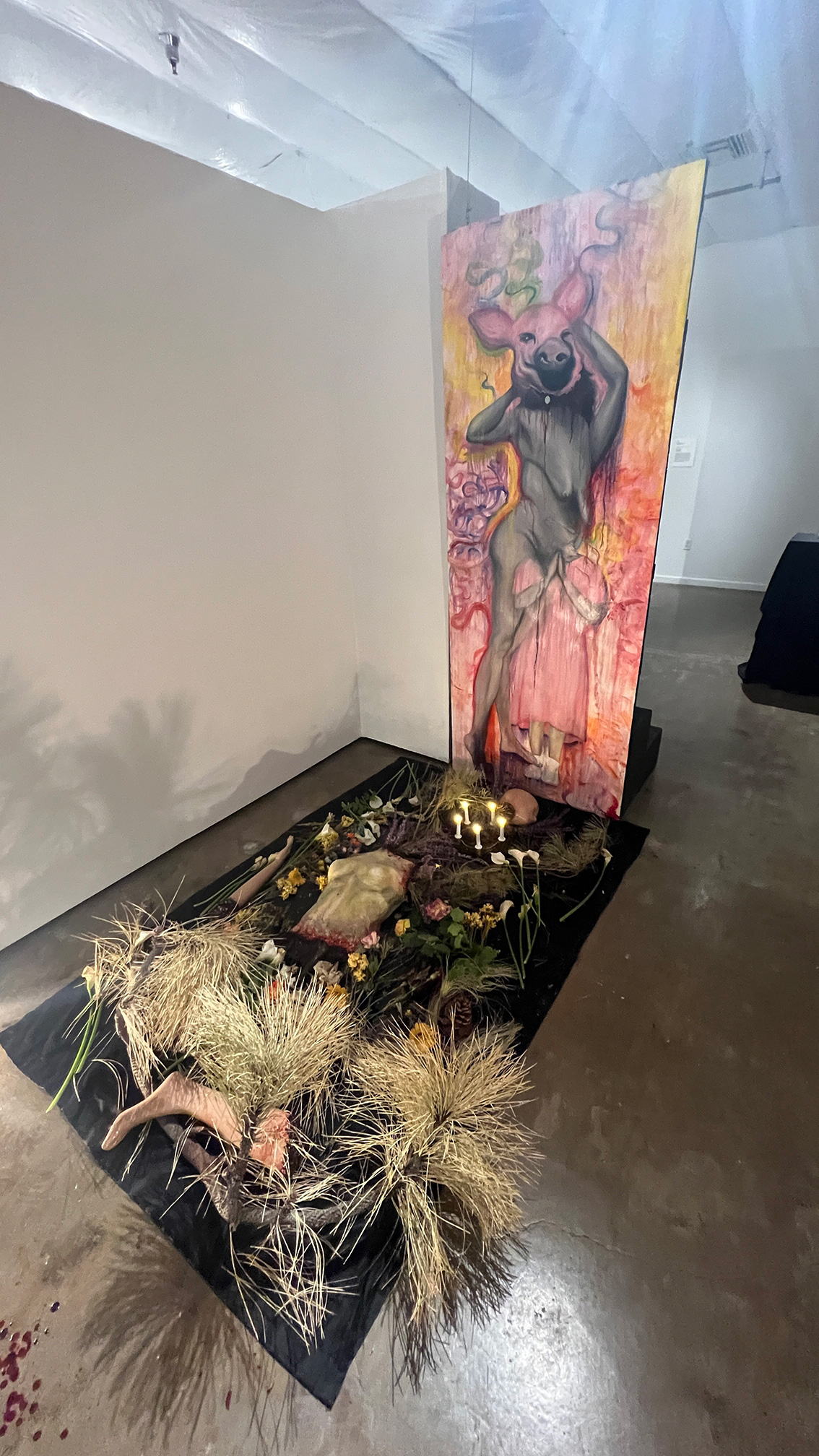 A large canvas has a painting of a pig-headed woman, with various items included food, mutilated body parts, and dead pine needles on the ground in front. There is a hole in the canvas which allows individuals to view to the piece through itself and on a mirror.