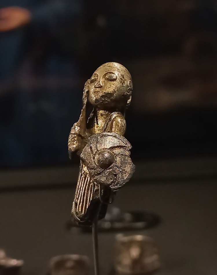 A small silver figurine of a woman with her hair tied back in a pleated skirt. She holds a shield in her left hand and a sword in her right.