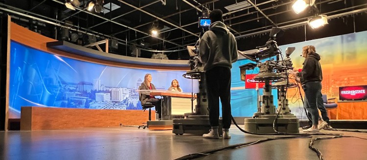The Fresno State Focus TV studio with two students operating large television cameras and two female anchors at the anchor desk on set.
