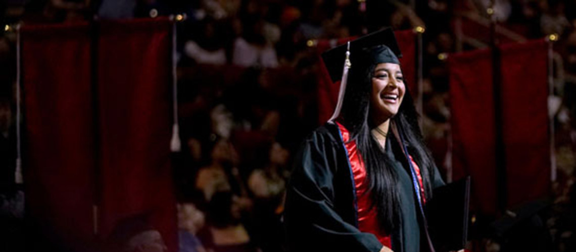 A Fresno State Graduate in a black cap and gown with a red and blue sash is illuminated by lights during graduation.
