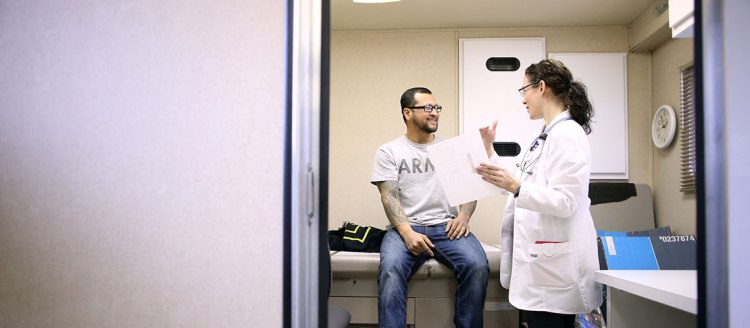 Male patient with a beard, glasses and a gray tshirt and jeans talks with medical a female professional in white scrubs holding a piece of paper in an exam room.