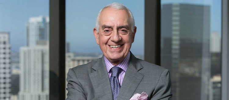 Ken Khachigian stands in front of a window overlooking city skyscrapers in a grey suit and purple tie.