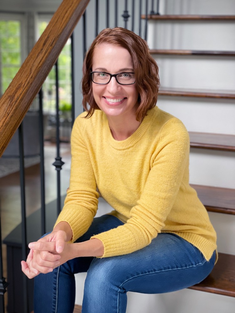Tiffany Crum in a yellow shirt and blue jeans sits on wooden stairs.