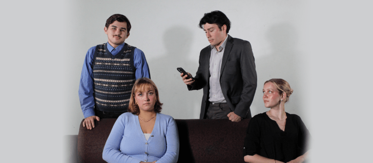 Two women sit on a couch and two men stand behind them -- one looking at his phone.