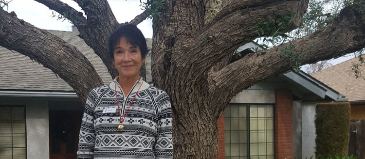 Carla Millar stands in front of a tree and a house in a white and black pattered sweater and a red string with a jingle bell around her neck.