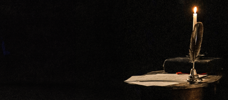A letter sits with a feather quill pen on a desk in the dark lit by a candle.
