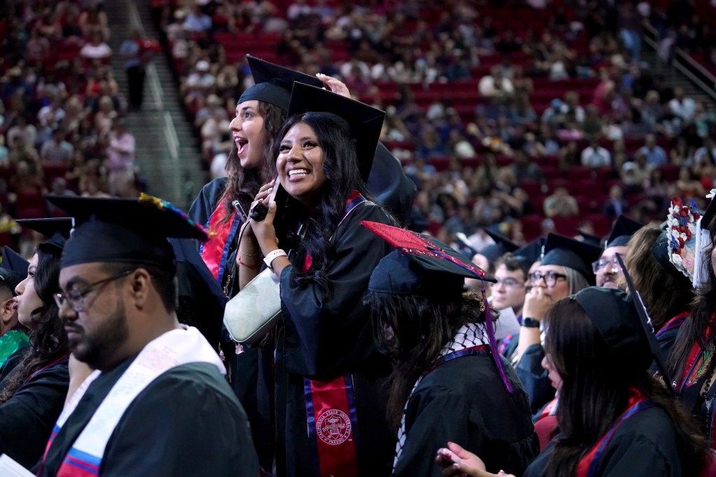 Two grads stand together. One is on the phone and looking at the audience.