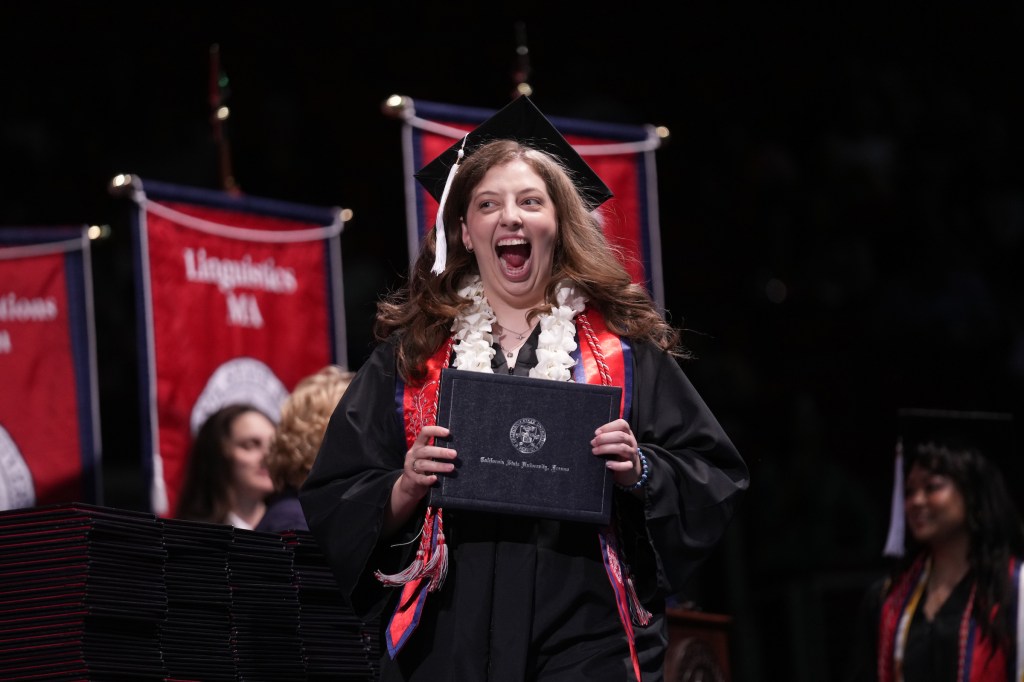 A graduate holds her degree on the stage while projecting overwhelming excitement.