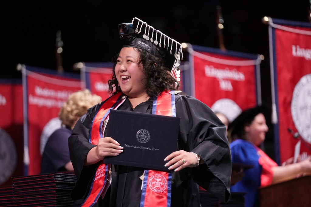 A grad with tassels hanging from her cap and a GoPro camera on her head holds her diploma as she crosses the stage.