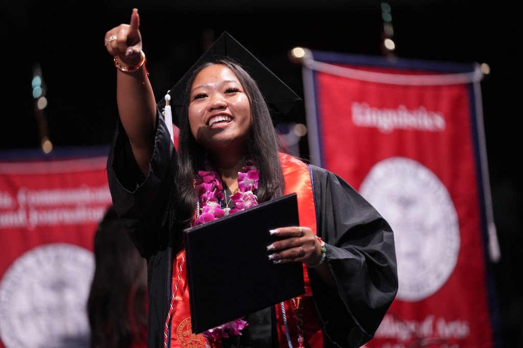 A grad point to someone in the crowd while holding their degree.