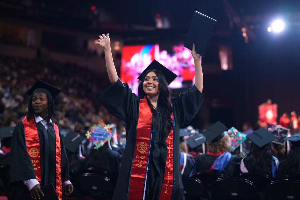 A graduate waves with both hands to the crowd. In one hand, is her degree.