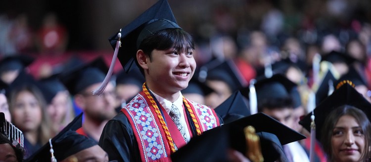 College of Arts and Humanities undergraduate Dean's Medalist Joshua Lor stands among sitting graduates in his regalia.