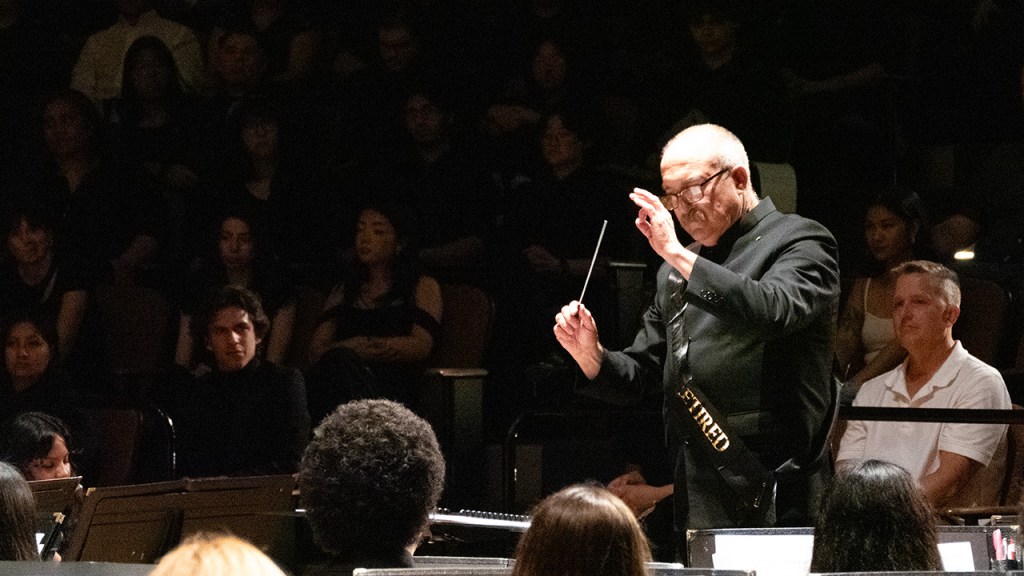 Dr. Gary Gilroy, dressed in black with a sash that says "the legend has retired" in gold letters, directs the final song of his final concert.