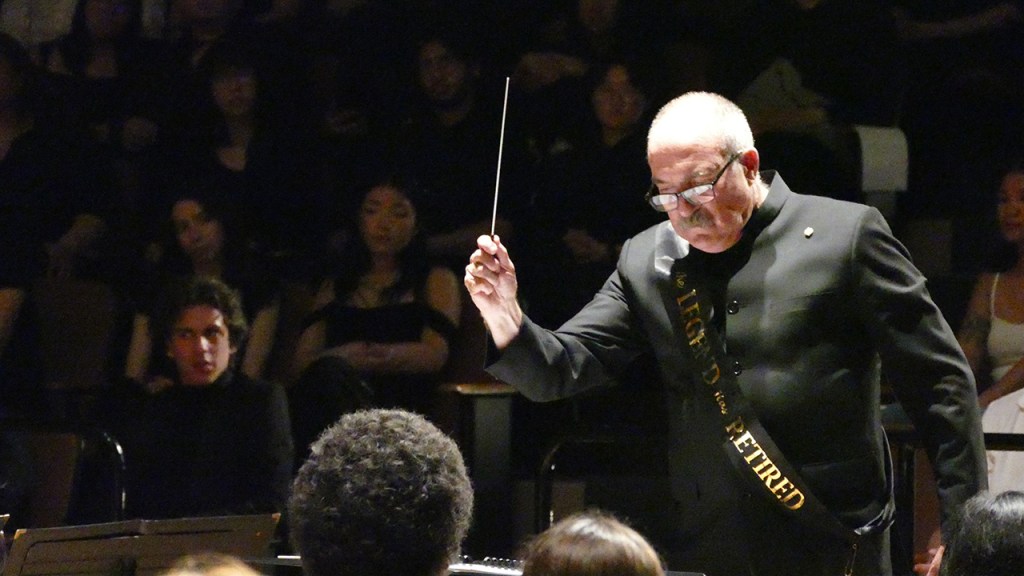 Dr. Gary Gilroy, dressed in black with a sash that says "the legend has retired" in gold letters, directs the final song of his final concert.
